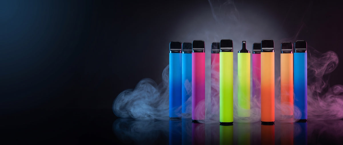 Colourful disposable vapes lined up in vapour on a dark reflective surface